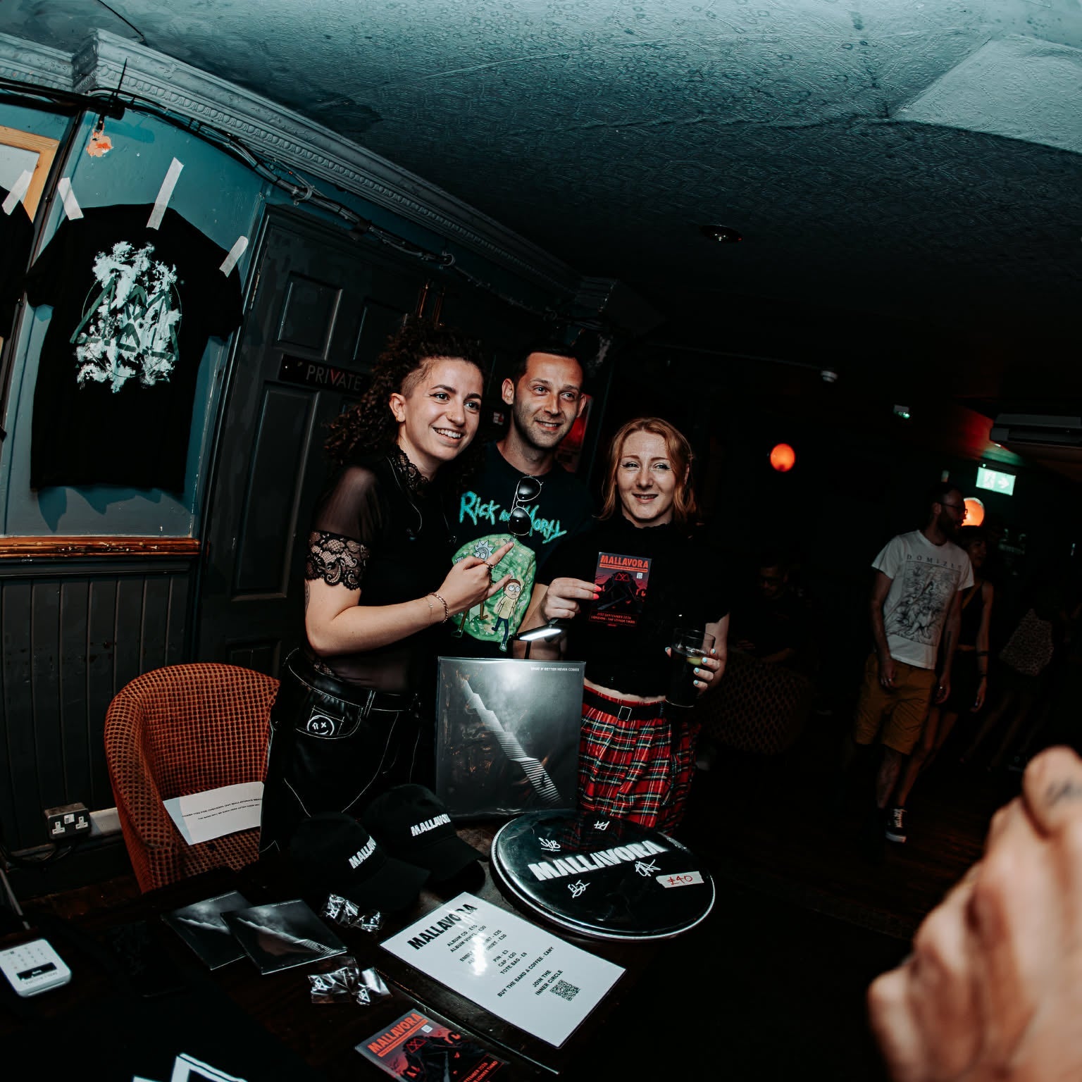 Three people standing together in a dimly lit venue, smiling and holding merchandise. Black graphic T-shirts with 'MALLAVORA' are visible on display, along with a table holding CDs and printed materials.