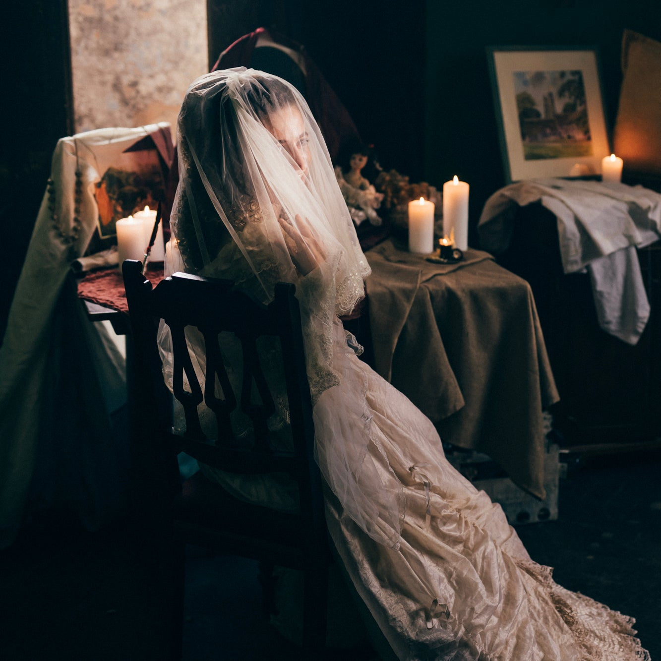 A woman in a long white wedding dress and veil sits in a dark room with candles, draped fabrics, and framed art in the background.
